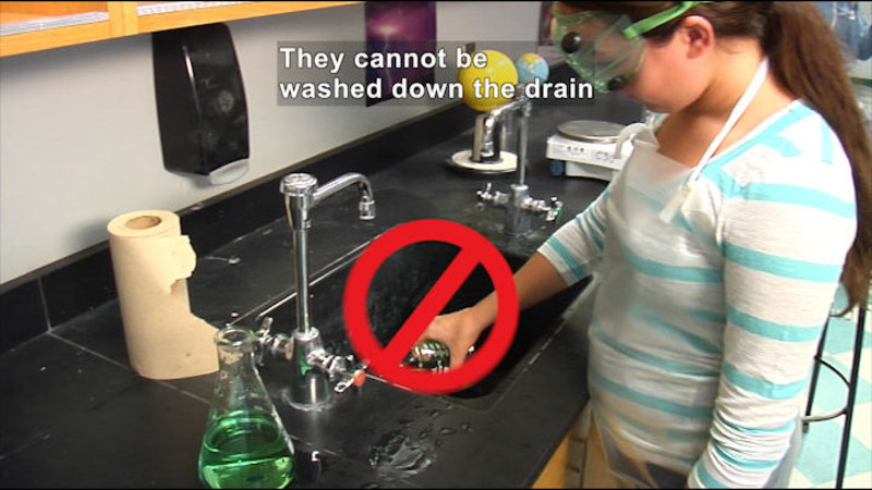 Person wearing safety goggles and pouring something down the drain of a sink. A red circle with a line through it covers the container being poured down the drain. Caption: They cannot be washed down the drain