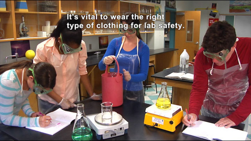 People wearing rubber gloves, plastic aprons, and protective goggles in a science classroom. Caption: It's vital to wear the right type of clothing for lab safety.