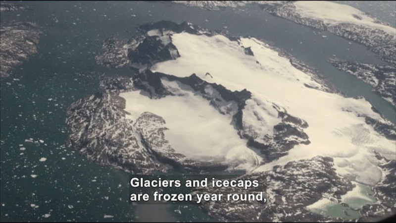 Aerial view of snow-and-ice-covered rock surrounded by icy water. Caption: Glaciers and icecaps are frozen year round,