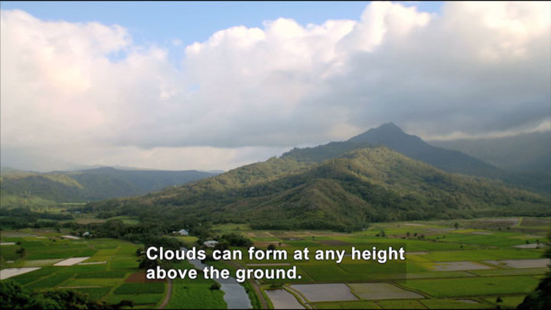 Green patchwork fields with a mountain rising in the background. Clouds cover most of the blue sky. Caption: Clouds can form at any height above the ground.