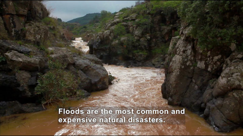 Muddy water rushing through a rocky channel. Caption: Floods are the most common and expensive natural disasters.