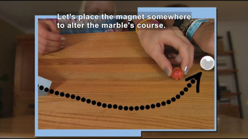 Person placing a magnet to one side of the path directly in front of a ramp. A dotted line projects the path of an object that will move from the ramp and curve toward the magnet. Caption: Let's place the magnet somewhere to alter the marble's course.