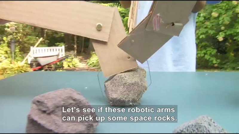 Cardboard pieces cut and attached together to resemble robotic arms attempting to pick up rocks. Caption: Let's see if these robotic arms can pick up some space rocks.