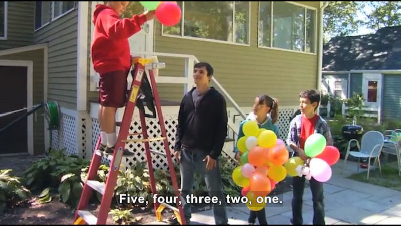 Four people in a yard holding balloons. One is on a ladder. Caption: Five, four, three, two, one.