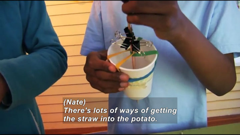 Person holding a paper cup with rubber bands and binder clips wrapped around a tubular object. Caption: (Nate) There's lots of ways of getting the straw into the potato.