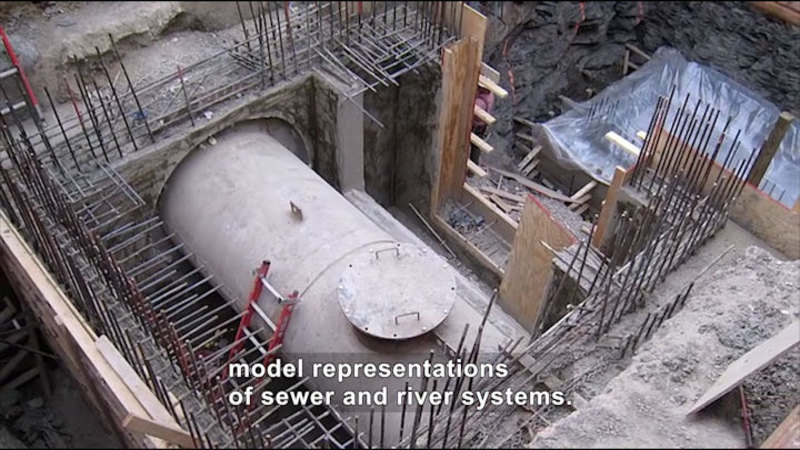 Seen from above, a large cylindrical tube with a hatch in a pit surrounded by wooden supports and metal bars. Caption: model representations of sewer and river systems.