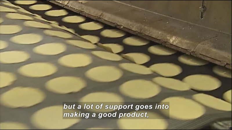A conveyor belt moves rows of light colored, flat, and round objects at a high speed. Caption: but a lot of support goes into making a good product,