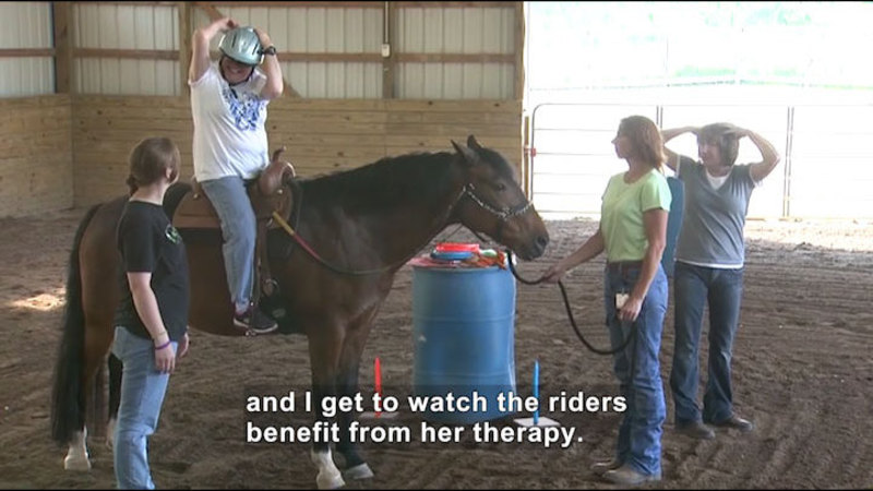 Three people stand around a horse. One holds the reins of the horse. A smiling person wearing a helmet is mounted on the horse. Caption: and I get to watch the riders benefit from her therapy.